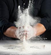 man in black uniform sprinkles white wheat flour in different directions, product scatters dust, black background, man sitting at a table