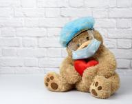 brown teddy bear sits in protective plastic glasses, a medical disposable mask and a blue cap against a background of a white brick wall. Protective accessories from the virus during an epidemic