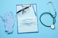disposable medical mask, plastic goggles and stethoscope on blue background, top view