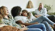 Laughing daughter embracing mother while sitting on couch with family in living room