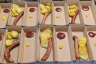 Large group of sausages served in takeaway food containers with fried potatoes tomato sauce and mustard sauce