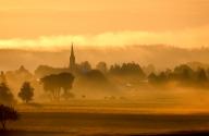 19 May 2024, Baden-Württemberg, Uttenweiler: The rising sun colors the fog around the church of St. Ursula in Dieterskirch yellow in the morning Photo: Thomas Warnack\/dpa