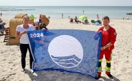 24 May 2024, Mecklenburg-Western Pomerania, Wustrow: Katrin Fischer (l), spa director of Wustrow, and Jan Schubert, lifeguard of the DLRG local group Fischland, present the "Blue Flag" 2024 on the eastern beach, which was previously awarded to 27 bathing areas and five marinas in Mecklenburg-Vorpommern. The "Blue Flag" has been awarded annually since 1987 in cooperation with the Foundation for Environmental Education (F.E.E.) and confirms special achievements in the field of environmental protection and sustainability. Wustrow on the Baltic Sea peninsula of Fischland is receiving this award for the 25th time this year. Photo: Bernd Wüstneck/dpa