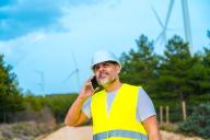Smiling worker in reflective waistcoat and helmet using phone in a wind energy