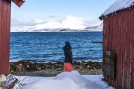 A person looks at the sea between two huts, surrounded by snow-capped mountains, Norway