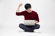 Young asian man feeling happy holding laptop computer while sitting on a floor isolated over white background