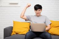 Young Asian man is browsing at his laptop and raising his arm up with celebrating success on sofa at home