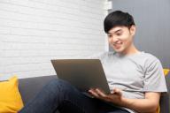 Young Asian man is browsing at his laptop and sitting on sofa at home
