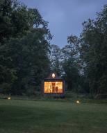 The Apiary pavilion seen at dusk from across a field, surrounded by trees. Interior lights are golden showing back of building, seating pits and exhibition space. The Beezantium at The Newt, Bruton, United Kingdom. Architect: Invisible Studio, 2021