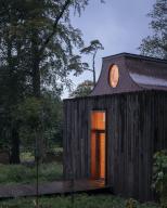 The Apiary pavilion seen at dusk, surrounded by trees. Interior lights are golden showing atrium window and entrance. The Beezantium at The Newt, Bruton, United Kingdom. Architect: Invisible Studio, 2021